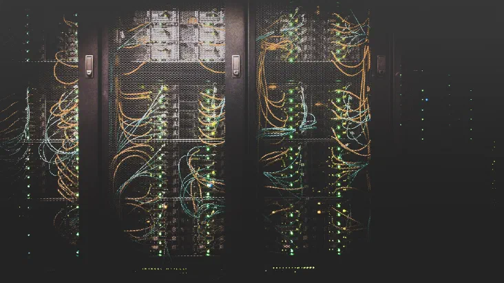 Rows of server racks with mesh doors, behind which orange and teal network cables loop between switches in a dim data center