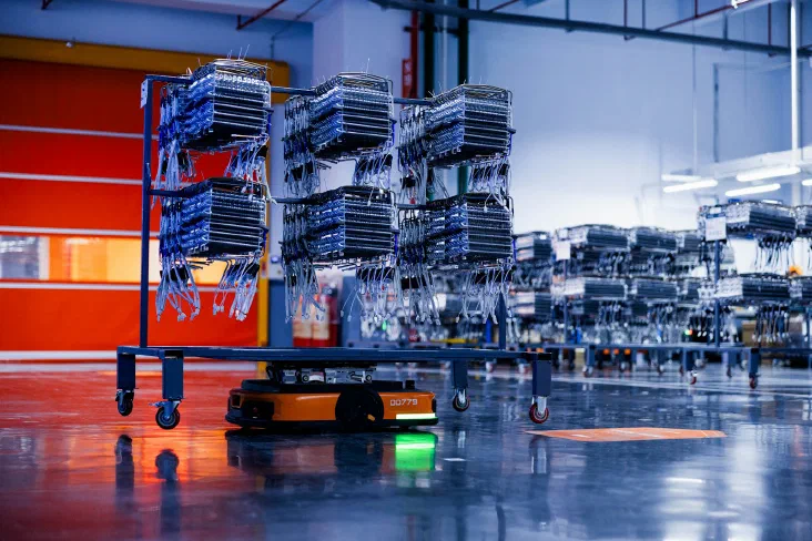 An automated guided vehicle carrying stacked racks of components through a factory warehouse, with more loaded racks lined up in the background