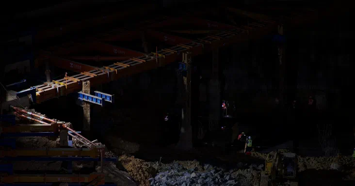 Eine Brücke im nächtlichen Bau, beleuchtet von Arbeitslichtern, mit Bauarbeitern in hochsichtbarer Schutzkleidung und einem Bagger darunter