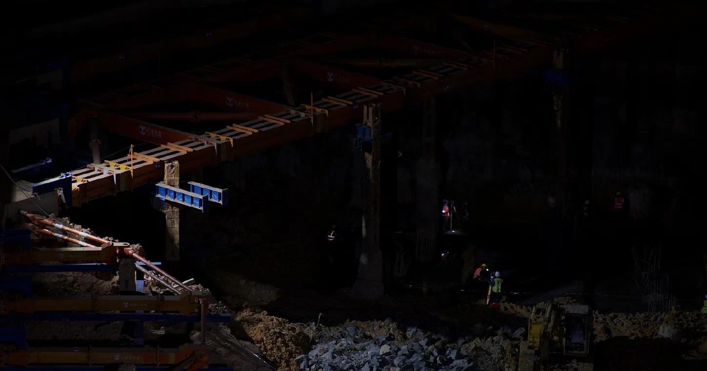Eine Brücke im nächtlichen Bau, beleuchtet von Arbeitslichtern, mit Bauarbeitern in hochsichtbarer Schutzkleidung und einem Bagger darunter